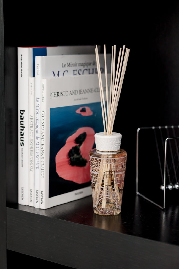 A shelf with books on art and design, such as Bauhaus and Christo and Jeanne-Claude, sits elegantly on a black surface. Accompanying them is the Baobab - My First Baobab Paris 8.45oz Reed Diffuser, with its transparent liquid and wooden sticks adding sophistication.