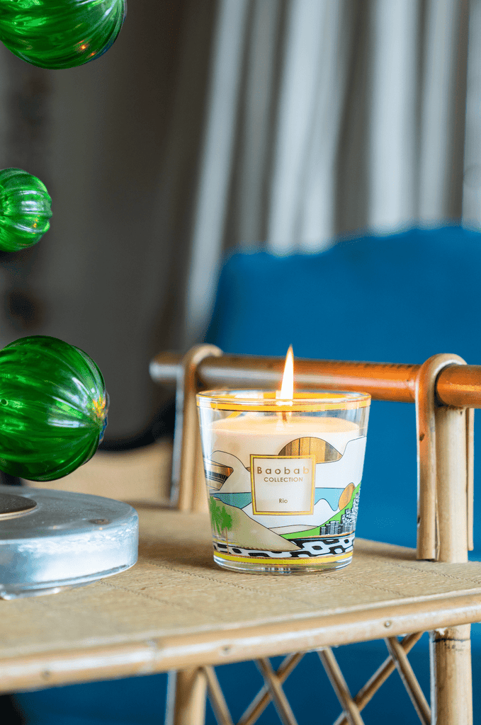 A Boabab - My First Baobab Rio 6.7oz Candle by Baobab with a colorful glass design sits on a wicker table, surrounded by green glass orbs and exuding a bergamot green tea musk aroma. A blue cushion can be seen in the blurred background.