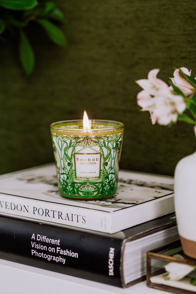 A green ornate glass Boabab - My First Baobab Tomorrowland 6.7oz Candle by Baobab sits on books, A Different Vision on Fashion Photography and Vogue, with a plant and pink flowers against a green fabric wall, crafting an elegant ambiance.