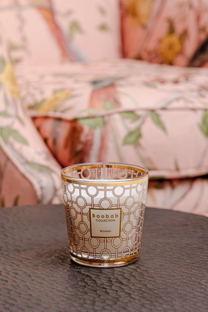 A Boabab - My First Baobab Womens 6.7oz Candle rests on a textured black table. This Baobab candle, with its gold and white geometric design, complements the pink floral-patterned armchair nearby, creating an elegant ambiance similar to rose-scented gifts.