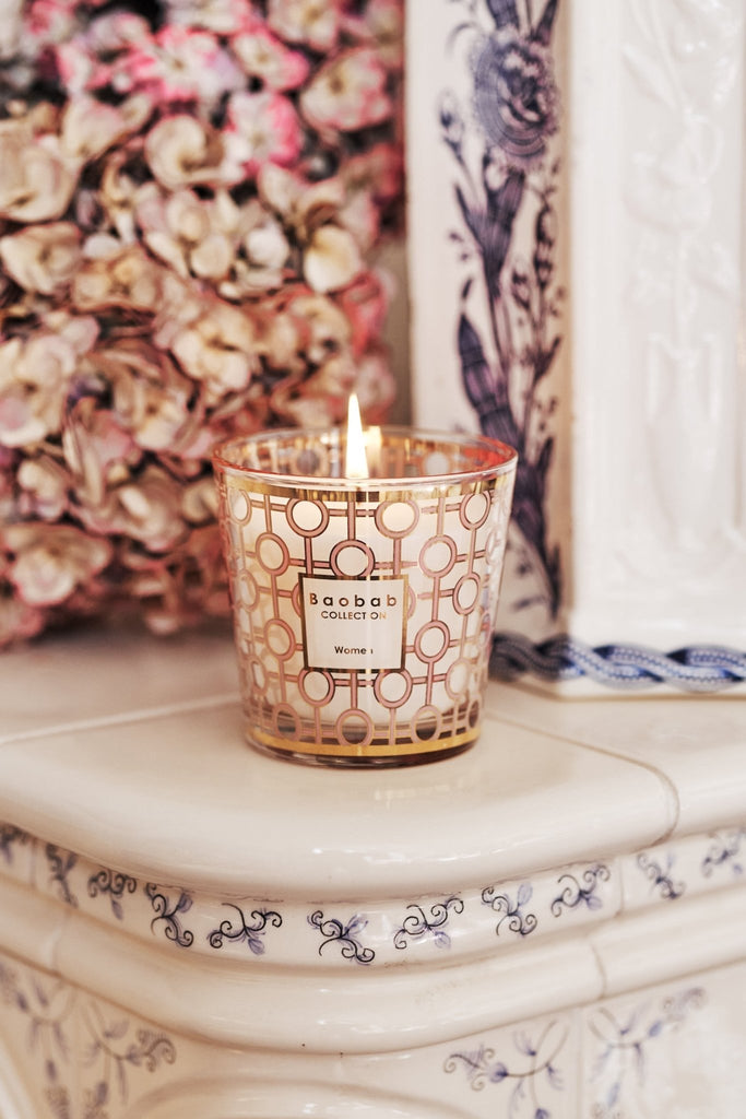 The Baobab - My First Baobab Womens 6.7oz Candle sits in an ornate glass holder on a white ceramic surface with blue floral patterns, complemented by a bouquet of pink and beige hydrangeas that enhance the elegant geometric gold design.