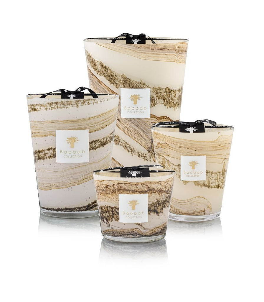 Four mouth-blown glass candles with marble-like brown and beige swirls are displayed, labeled Baobab - Sands - Siloli Max 10 Candle. The cedarwood scent from the Baobab brand fills the air as they are pyramid-stacked with ribbons.