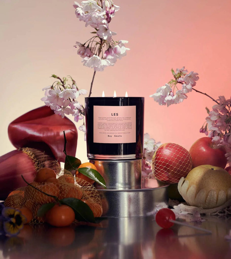 The Boy Smells - Ash 27oz Candle rests on a metallic stand, surrounded by apples, pears, and citrus with decorative pink blossoms above. Made from coconut and beeswax and infused with a woodsy scent, it is softly lit in warm colors for a cozy atmosphere.