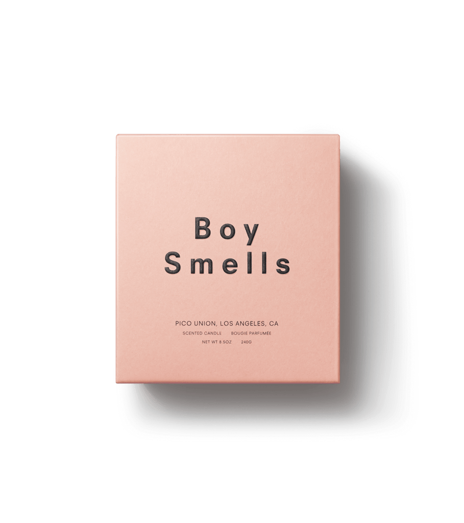 A square pink box displays Boy Smells in black, with Pico Union, Los Angeles, CA, Scented Candle, and weight details. Set against a plain black backdrop, the packaging suggests the smoky woody essence of rare woods.