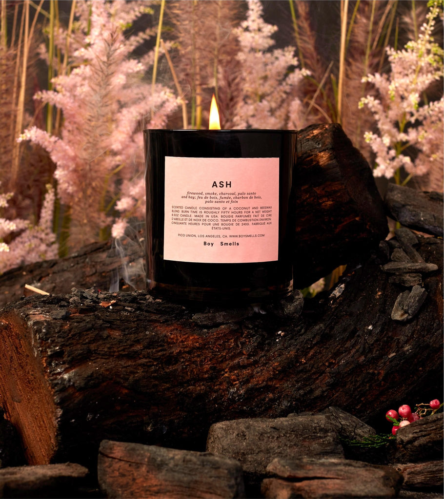 The Boy Smells - Ash 8.5oz Candle in a black container rests on charred wood with pink foliage, offering an inviting atmosphere enriched by rare woods.