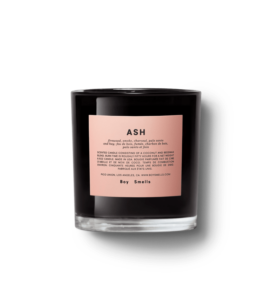 The Boy Smells - Ash 8.5oz Candle features smoky woody notes of firewood, smoke, black charcoal, and palo santo. Its sleek black glass design boasts a light pink label with black text, offering an elegant contrast against the shiny finish.