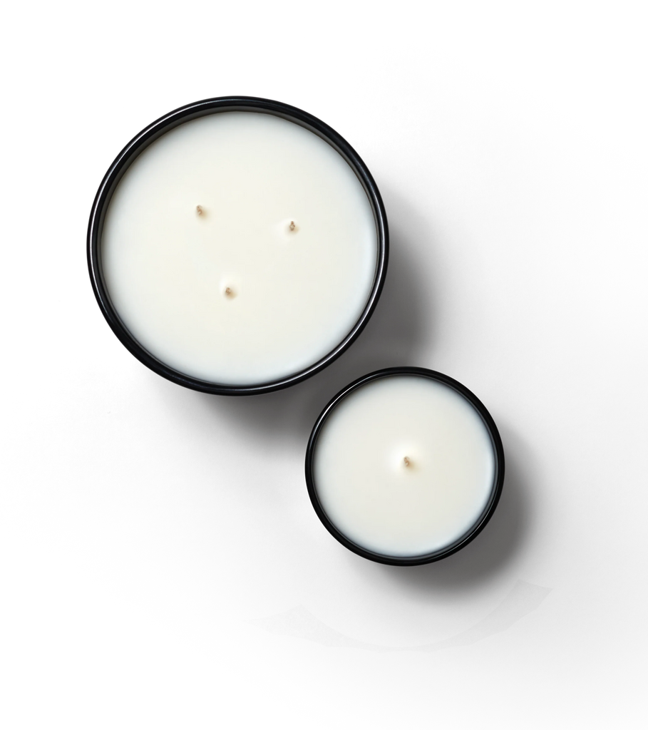 Against a black background, two Boy Smells candles in black containers are displayed. The larger Cedar Stack scented candle, crafted with coconut wax and featuring three wicks, is positioned above the smaller one-wick candle.