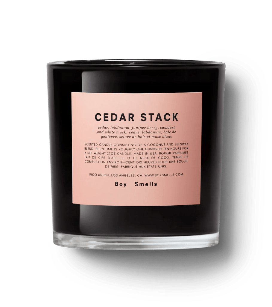 The Boy Smells Cedar Stack 27oz Candle, featuring a black exterior and a pink label reading Cedar Stack, emits a woody scent with cedar, labdanum, and juniper berry notes. Crafted from coconut wax, it elegantly showcases the Boy Smells brand at the bottom of its label.