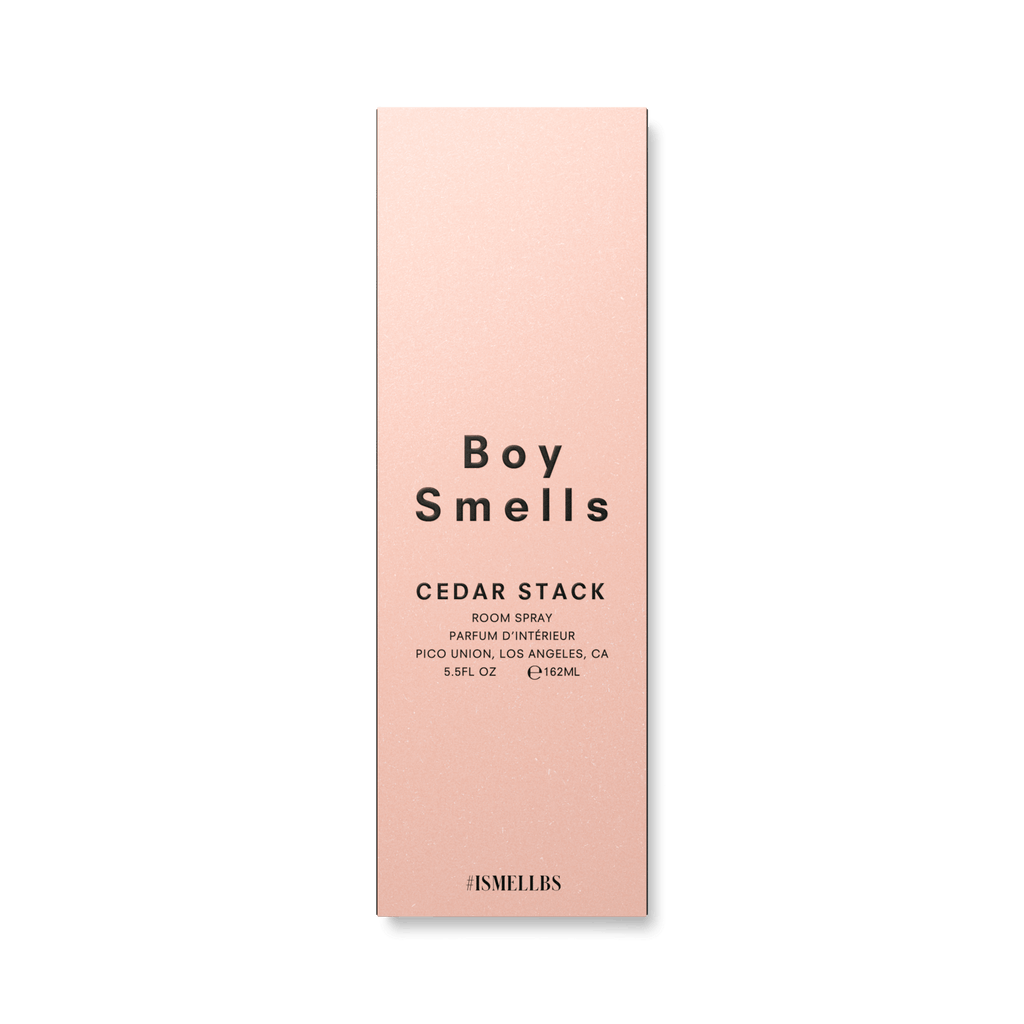 A pink rectangular box with bold black letters displays Boy Smells, featuring CEDAR STACK and ROOM SPRAY below, indicating a captivating wood scent. Additional details such as location, volume (5.5oz), and hashtag #SMELLBS are set against a stark black background.