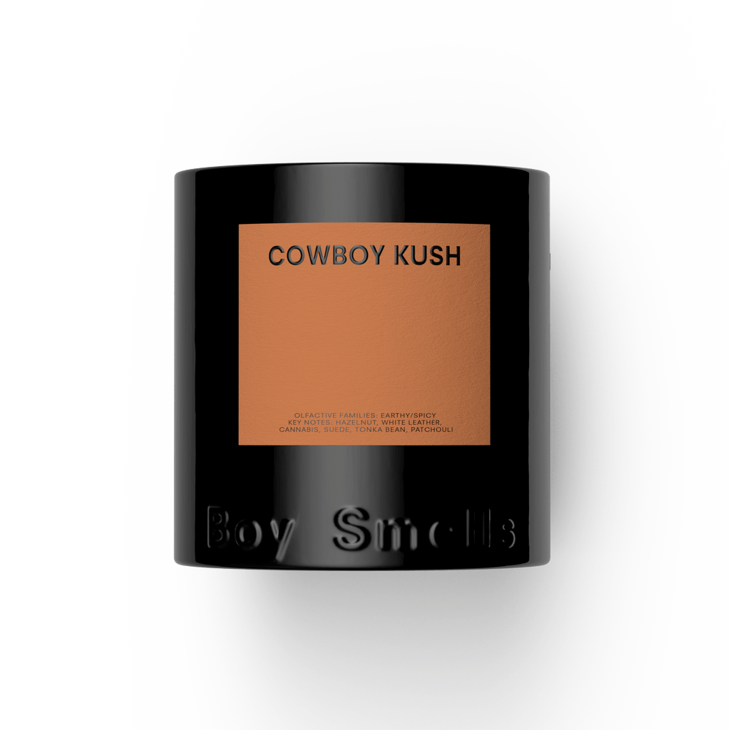A 27oz Boy Smells - Cowboy Cush candle in a black jar with a tan label, featuring earthy and spicy scent notes. "Boy Smells" is embossed on the lower part of the jar. The background is plain white.