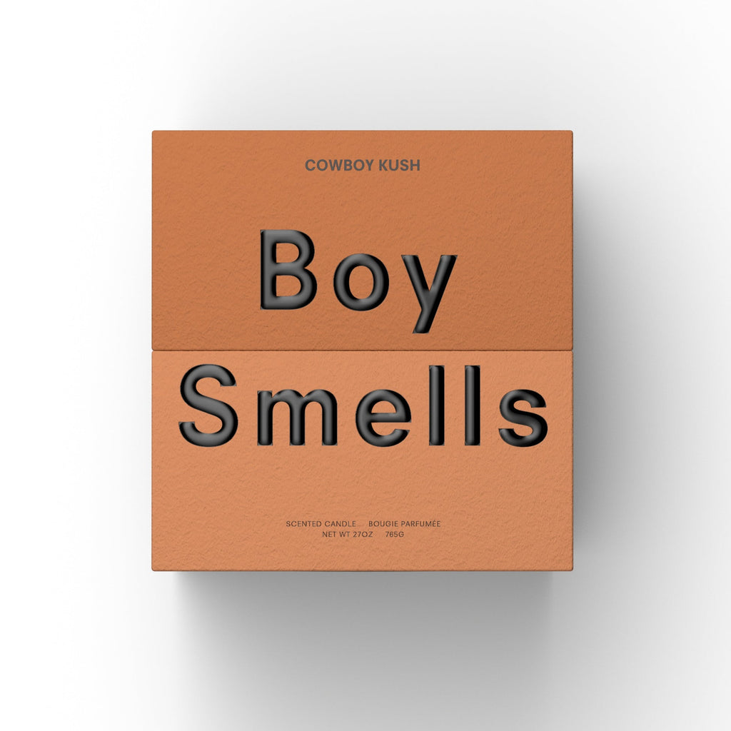 A square, terracotta box features "COWBOY CUSH" above bold black "Boy Smells" text, with smaller print noting its earthy, spicy scent in the 27oz Boy Smells - Cowboy Cush Candle.