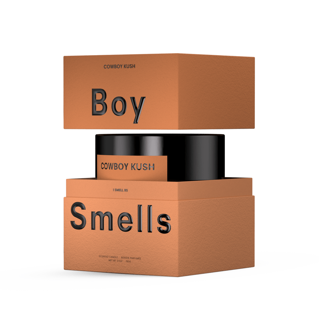 A product display shows a 27oz Boy Smells - Cowboy Cush candle in a black jar, with earthy spicy scent, placed between two orange-brown Boy Smells boxes labeled “Boy” and “Smells” in bold black text on a white background.