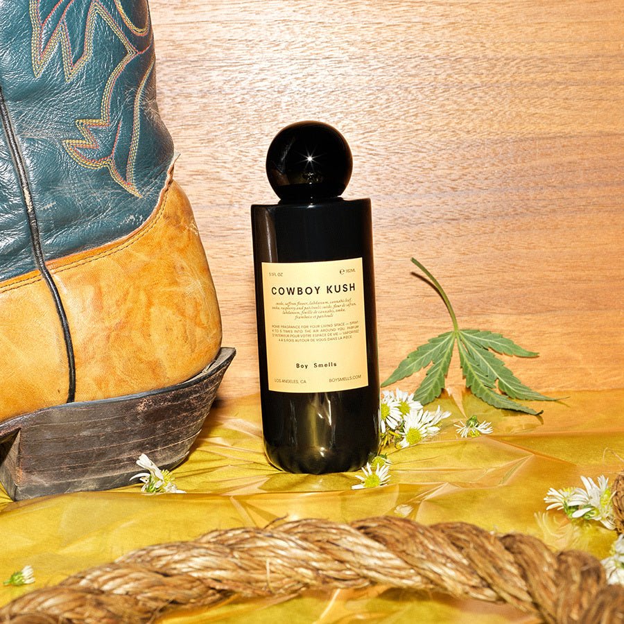 A Boy Smells - Cowboy Kush 5.5oz Room Spray sits on wood next to a blue and yellow cowboy boot, surrounded by an earthy/spicy scent with a hemp leaf, small white flowers, and braided rope enhancing the rustic vibe.