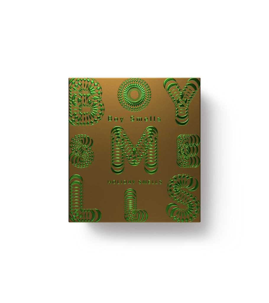 A box with a golden background and intricate green patterns evokes chai warmth. The design forms Boy Smells, arranged symmetrically, exuding holiday serenity. It casts a shadow to the right. Boy Smells - Holiday Smells 8.5oz Candle by Boy Smells.