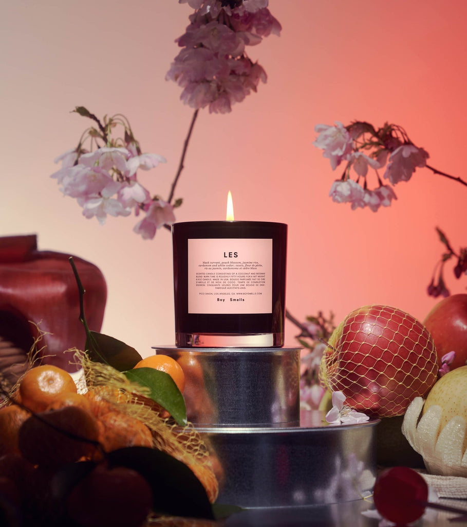 A Boy Smells - LES 8.5oz Candle sits on a reflective surface, surrounded by cherries, oranges, apples in netting, and delicate pink flowers above, creating a fruity/floral ambiance against a warm reddish hue.