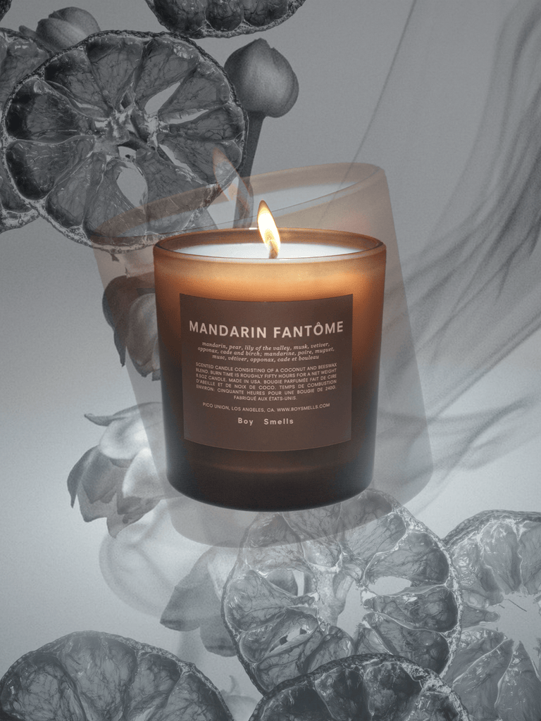 The Boy Smells - Mandarin Fantôme 8.5oz Candle in a frosted glass jar casts a warm glow, accented by halved and whole mandarins on branches against a grayscale backdrop, with delicate smoke wisps evoking smoked mandarin peels swirling around the candles earthy scent.