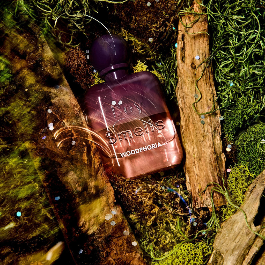 The Boy Smells - Woodphoria Eau de Parfum 50ml sits atop green moss and tree bark in a forest-inspired scene, with its purple glass bottle evoking fresh fig leaves and sandalwood notes.