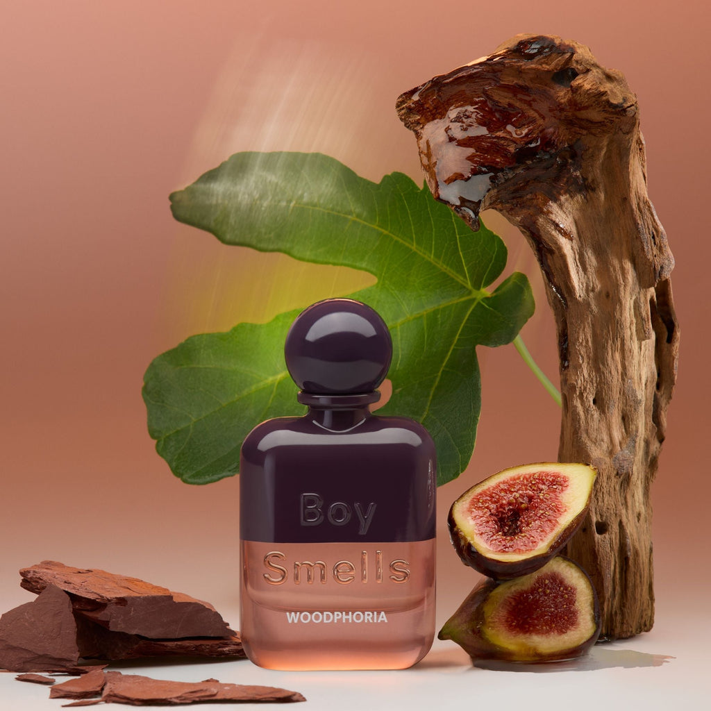 The Boy Smells - Woodphoria Eau de Parfum 50ml is showcased with fig leaves, a halved fig, wood pieces, and brown slate on a warm beige background, reflecting its earthy and fruity notes balanced by creamy sandalwood.