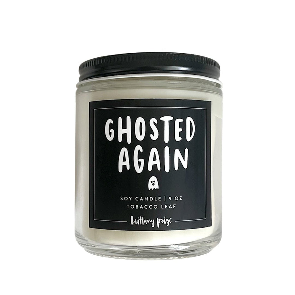 The Brittany Paige - Ghosted Again candle is a 9 oz white soy candle in a glass jar with a black lid, featuring the Ghosted Again label with a small ghost icon. It offers an aromatic tobacco leaf scent and is crafted from natural wax.