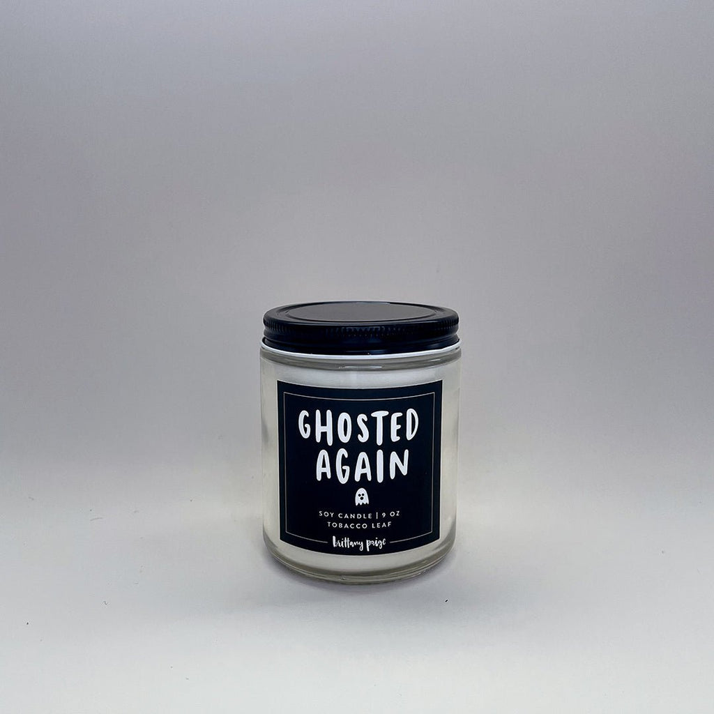 Against a plain background sits the Brittany Paige - Ghosted Again Candle, featuring a white jar with a black lid. The label, adorned with a ghost illustration, reads GHOSTED AGAIN and describes the rich Tobacco Leaf essence of this 9 oz soy candle made from natural wax.