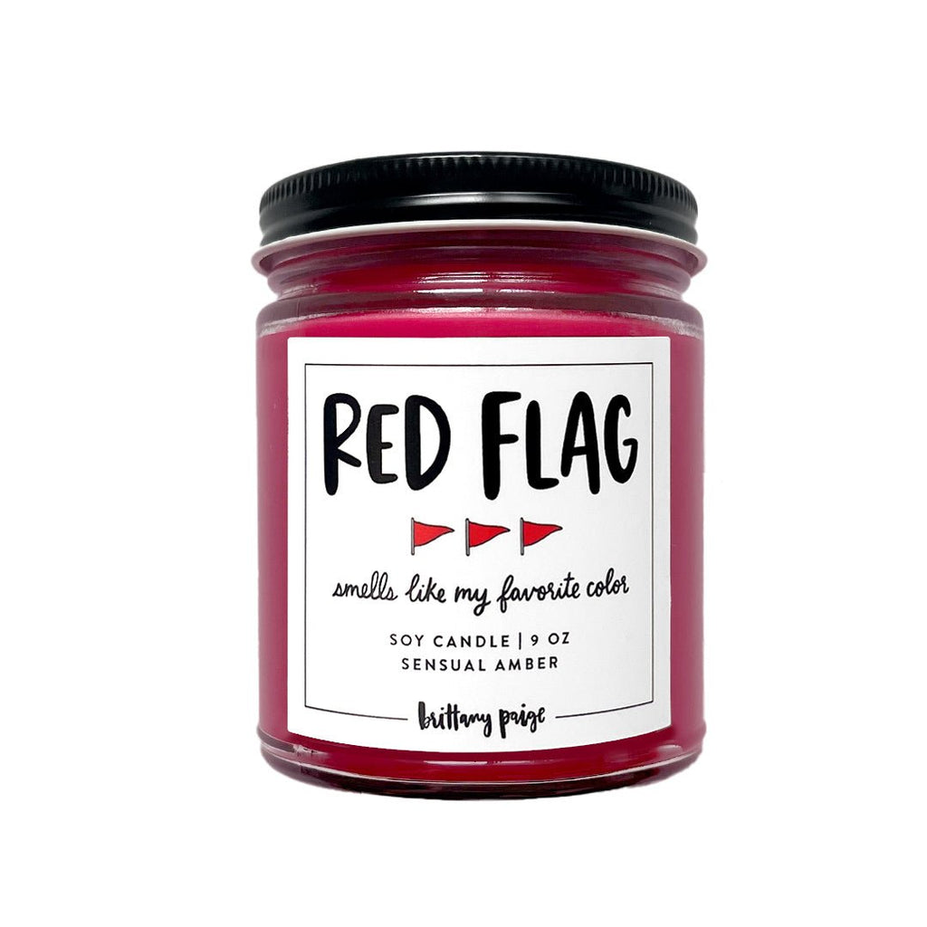 The Brittany Paige Red Flag candle features a sensual amber aroma, striking red hue, and sleek black lid. Adorned with tiny flag icons, the label reads smells like my favorite color. It burns for up to 50 hours.