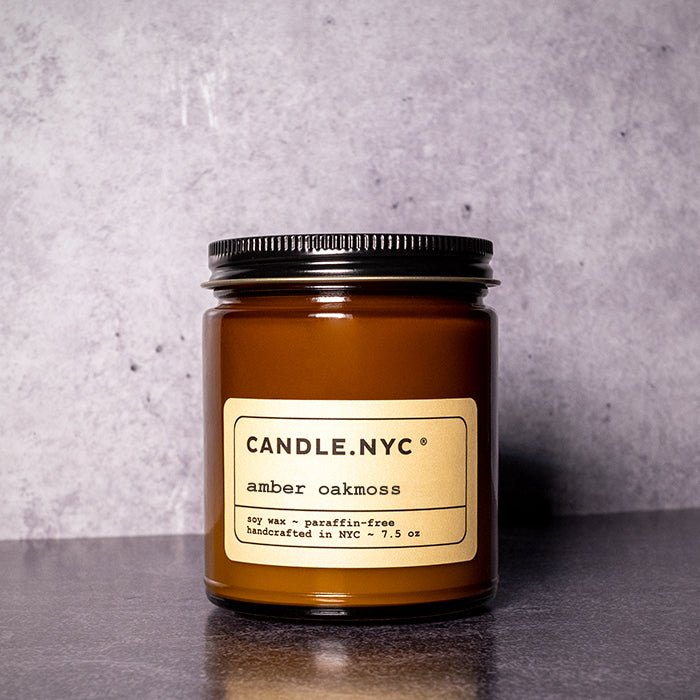 A Candle NYC - Amber Oakmoss candle from Candle.NYC sits against a gray backdrop, featuring an eco-friendly label that highlights its soy wax and paraffin-free composition. Handcrafted in NYC, it weighs 7.5 oz with a black screw-on lid topping the amber glass jar.