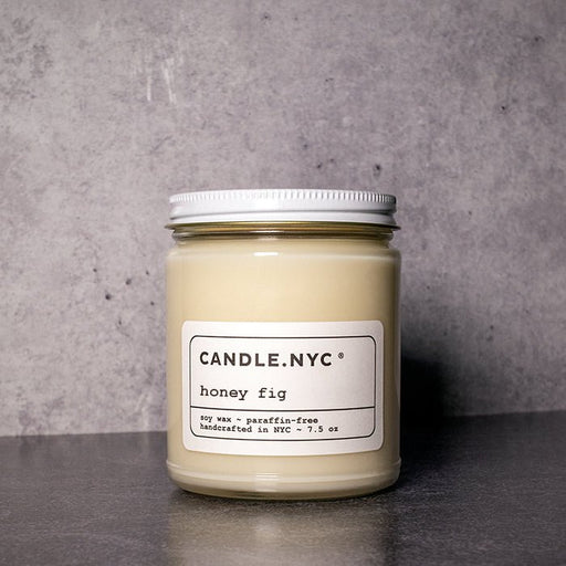 The Candle NYC - Honey Fig by Candle.NYC is a soy wax jar candle with a delightful honey fig fragrance, eco-friendly and paraffin-free, displayed elegantly against a light gray textured background.