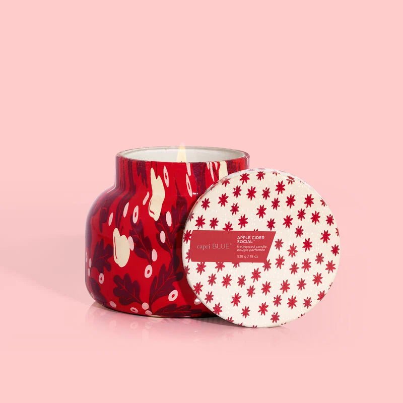 The Capri BLUE Apple Cider Social Holiday Pattern Play Signature Jar, an 8 oz candle, exudes a warm fragrance. Its red jar and lid feature abstract floral patterns with festive little red flowers on a light background, set against a soft pink backdrop for a cozy holiday ambiance.