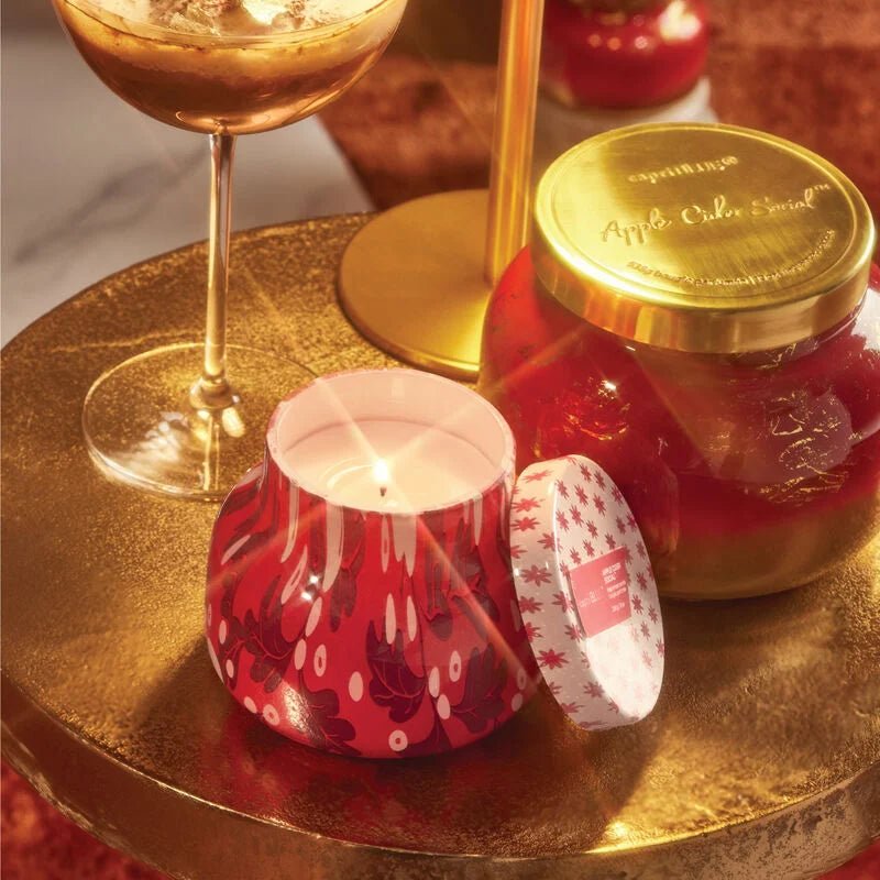 A Capri BLUE Apple Cider Social candle in a festive holiday holder emits a cozy scent, placed on a gold-patterned table with iced coffee and Apple Cider Spread. The plush red carpet adds warmth to the inviting atmosphere.