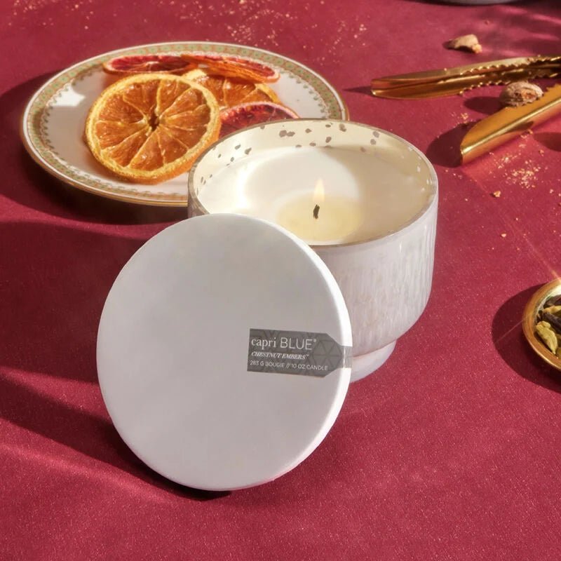 A warm, elegant scene features a lit Capri BLUE candle from their Chestnut Embers Holiday collection on a rich red cloth beside its cap. An inverted jar and a plate with dried orange slices and gold tongs complete the cozy arrangement.