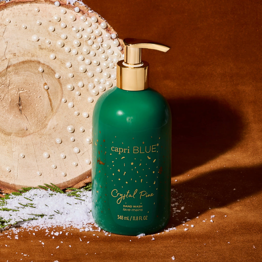 A green bottle of Capri BLUE - Crystal Pine Glimmer Hand Wash with gold text and a gold pump rests on a brown surface, surrounded by artificial snow, pine sprigs, and a wooden log adorned with white beads in the background.