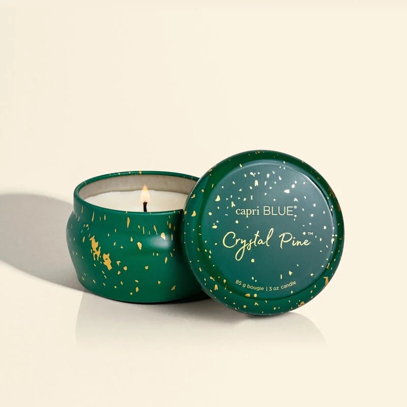 The Capri BLUE - Crystal Pine Glimmer Mini Tin 3 oz Candle in green emits a frosted fir aroma for a festive feel. Slightly open, the lid displays capri BLUE Crystal Pine in white with gold speckles, set against a plain, light background.