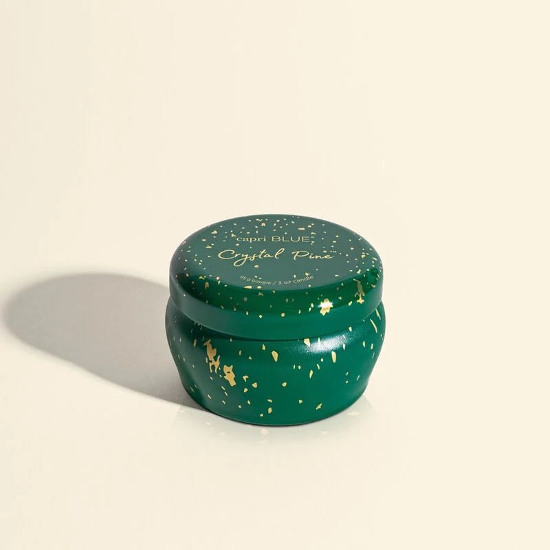 The Capri BLUE - Crystal Pine Glimmer Mini Tin 3 oz Candle is a small, round green container with gold speckles and Crystal Pine text on the lid. Ideal for stocking stuffers, it rests on a light background, casting a soft shadow and radiating a warm holiday atmosphere.