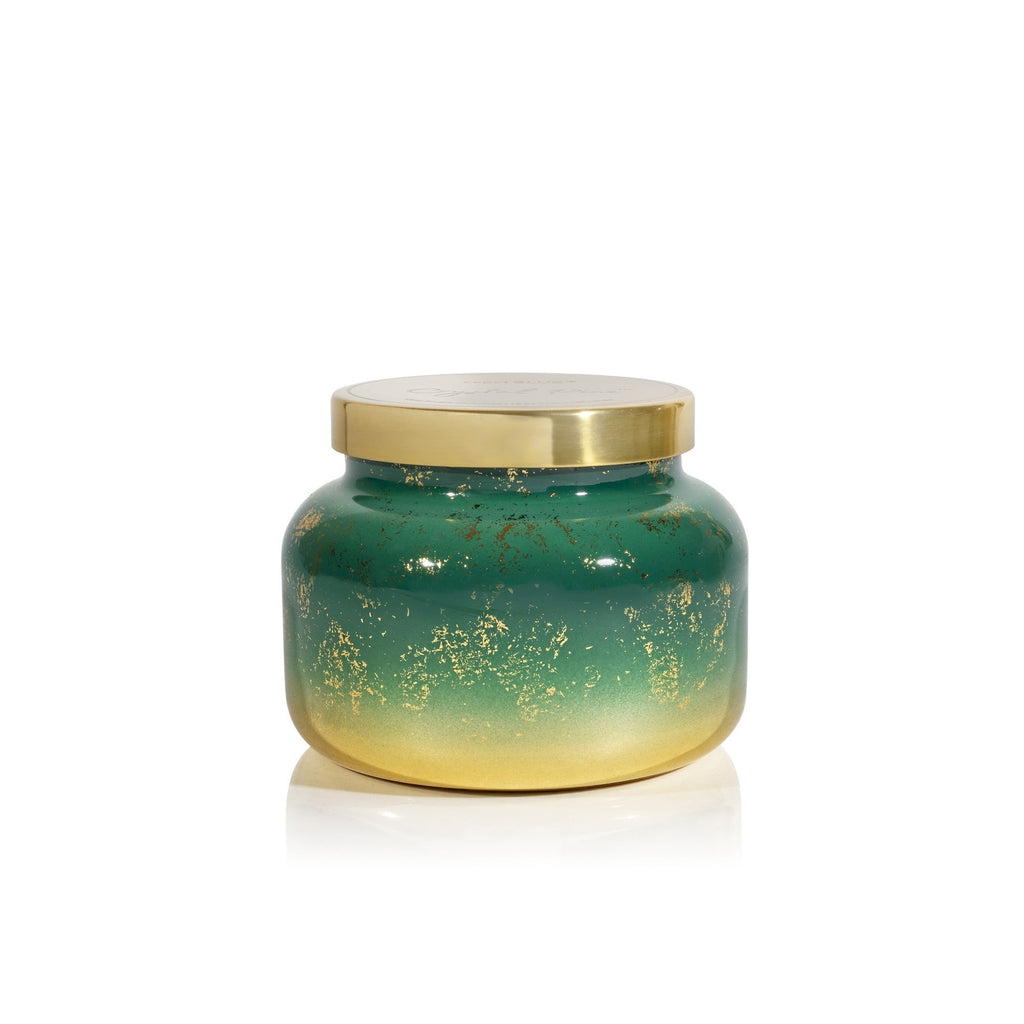 The Capri BLUE - Crystal Pine Glimmer Signature Candle, 19oz features a green jar fading to gold with gold flecks and a gold metal lid. This elegant winter candle by Capri BLUE is showcased against a plain white background.
