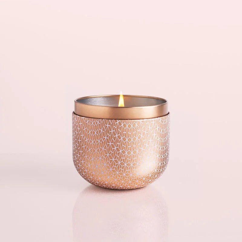 A Capri BLUE - Pink Grapefruit & Prosecco Gilded Tin 12.5 oz Candle sits on a light surface in a geometric-patterned tin holder, emitting a warm sheen. The small flame casts a soft glow with scents of pink grapefruit and prosecco.