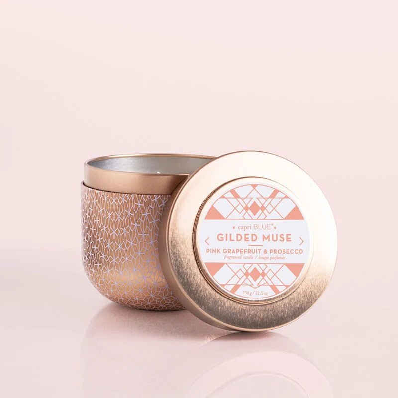 A candle jar with a gold geometric pattern sits with its open lid on a light pink surface. The lid reads Capri BLUE - Pink Grapefruit & Prosecco, indicating a fragrant scent. This 12.5 oz Gilded Tin candle boasts a metallic finish, adding elegance to any space.