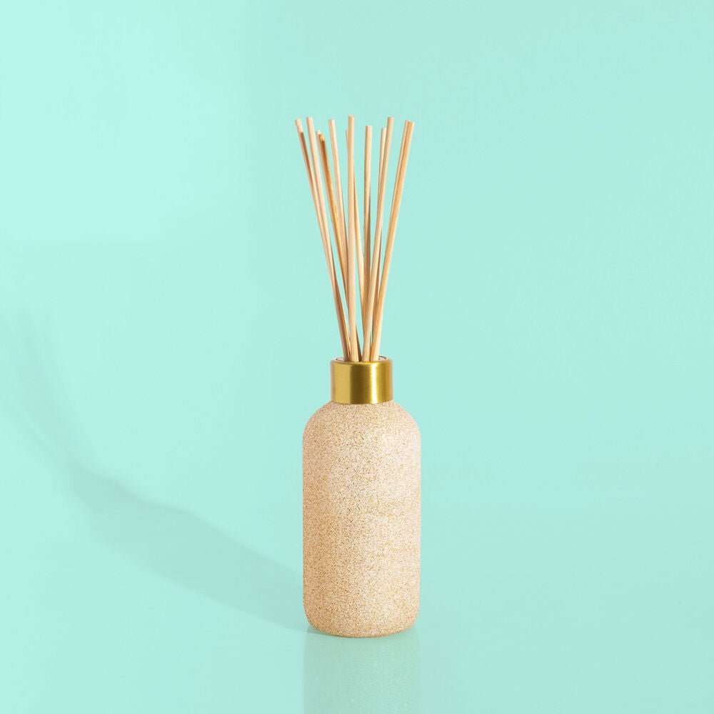 The Capri BLUE - Pumpkin Dulce 8oz Reed Diffuser, with a textured beige bottle and gold cap, features slender wooden reeds against a light turquoise backdrop. Ideal for an autumn scent or holiday gift, it casts a soft shadow on the shiny surface.