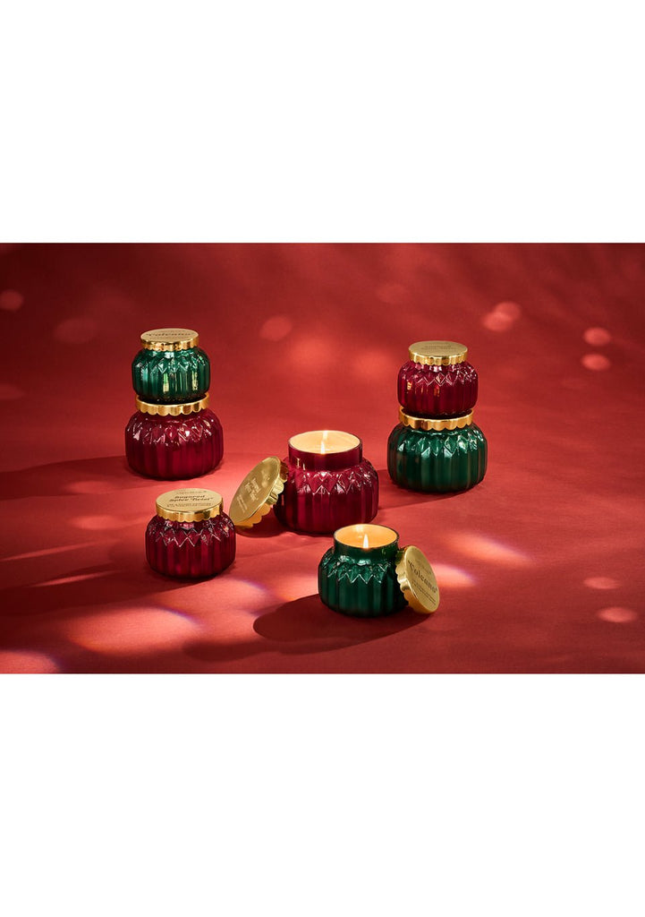 Several colorful, round Capri BLUE - Sugared Spice Twist Signature Candles (19oz) with gold lids are displayed on a red surface. Some are stacked, and two are open with flames burning inside, creating a warm and festive atmosphere.