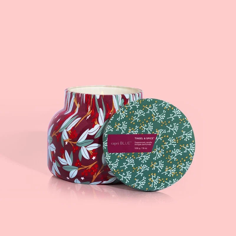 A 19 oz decorative candle in a signature red jar with white and green leaf patterns, featuring a green lid with small white floral designs. The label says Capri Blue Tinsel & Spice. Enjoy the Holiday Pattern Play against a solid pink background from Capri BLUE.