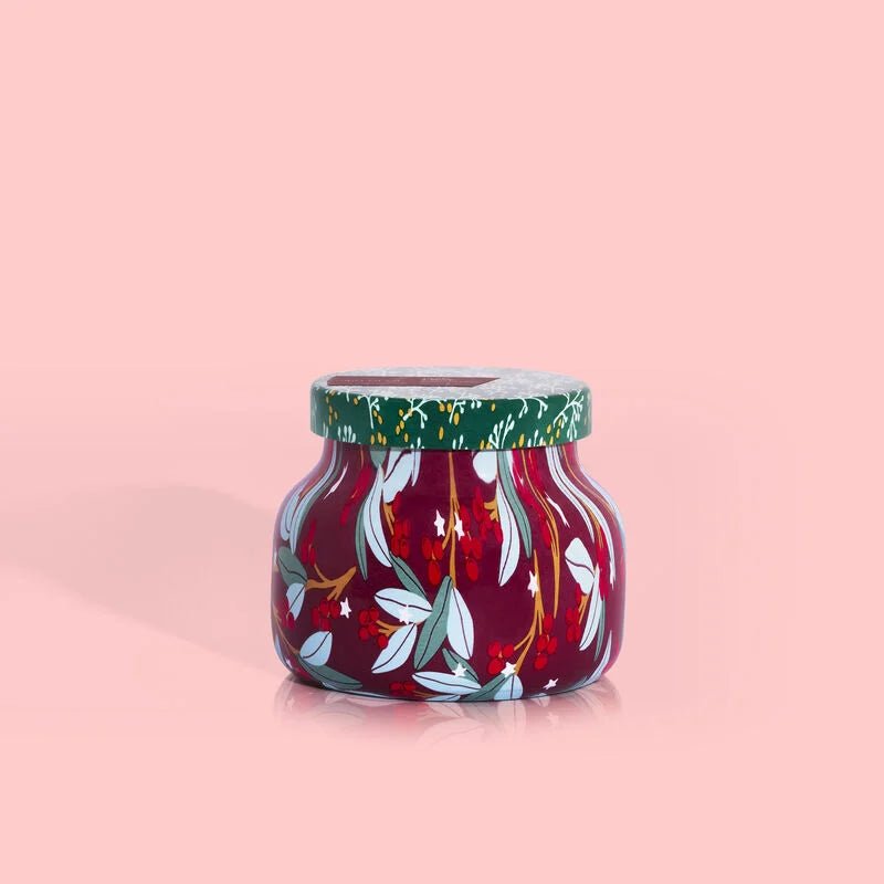 The Capri BLUE Tinsel & Spice Holiday Pattern Play Signature Jar 8 oz Candle features a floral leaf pattern on a red base with a green lid, capturing the festive spirit of the season and set against a plain pink background.