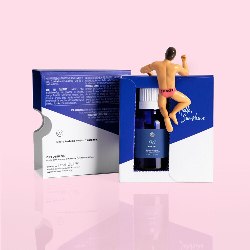 A blue box labeled Capri BLUE - Volcano 0.5oz Diffuser Oil features white text and suggests sugared citrus notes. A small figure in red swim trunks reading SPENCER rests on the box, set against a soft pink background that enhances the tropical feel.