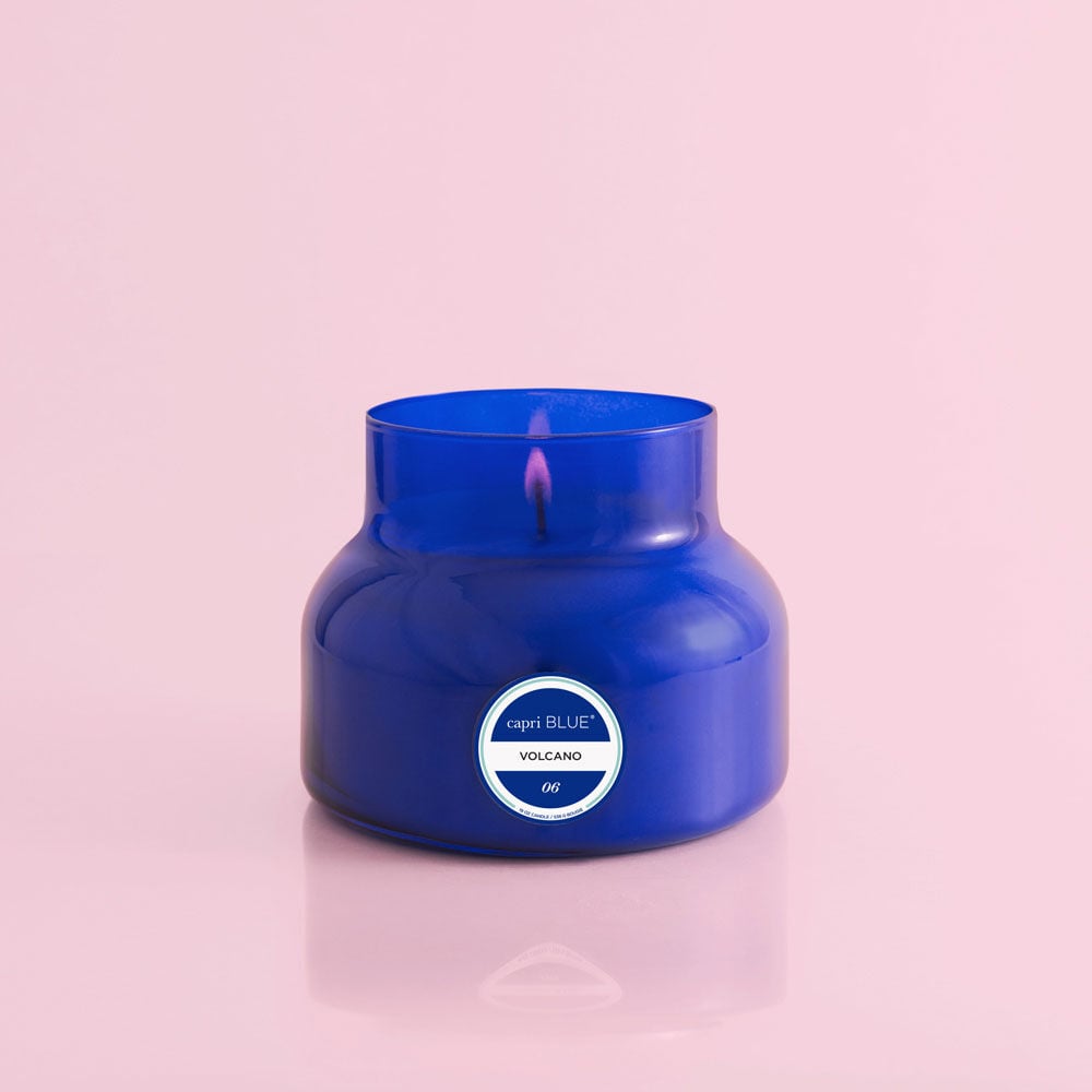 A Capri BLUE - Volcano 19oz Blue Signature Jar Candle with a single lit wick sits on a shiny pink surface against a pale pink background. The modern, rounded blue glass jar features a white label on the front.