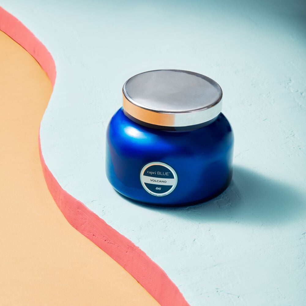 A Capri BLUE - Volcano 19oz Blue Signature Jar Candle, featuring a silver lid and the Capri BLUE brand label, rests on a textured pastel blue surface beside a curving peach and pink border.