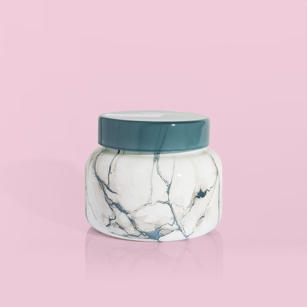 The Capri BLUE - Volcano 19oz Marble Signature Jar Candle, featuring gray marble veining and a matte gray lid, is shown against a pale pink background, suggesting its signature Volcano fragrance inside.
