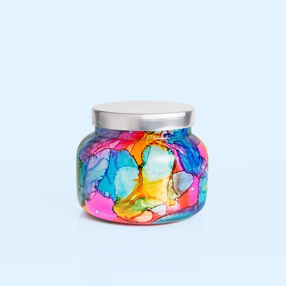 The Capri BLUE - Volcano 19oz Rainbow Watercolor Signature Jar Candle features a vibrant rainbow watercolor design and a silver lid, making it stand out beautifully against a light blue backdrop.
