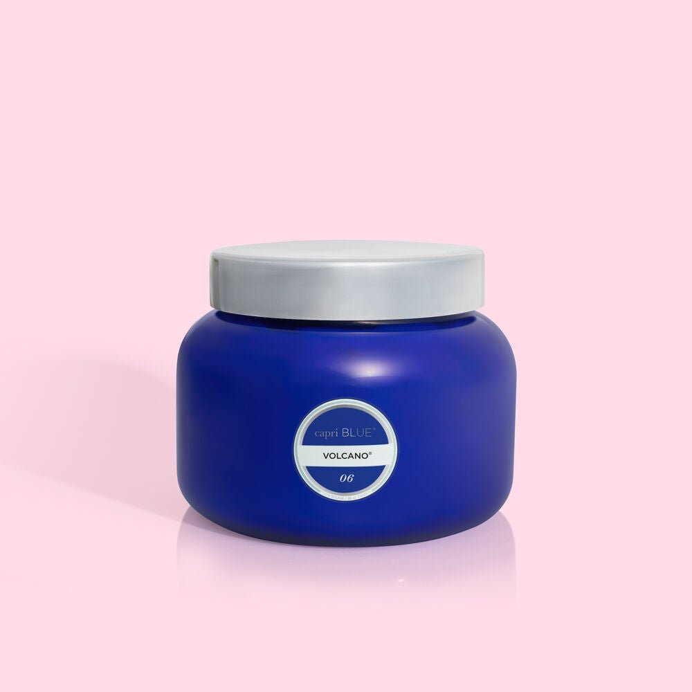 The Capri BLUE - Volcano 28oz Blue Oversized Jar Candle, with a silver lid and a sugared citrus scent, is displayed on a light pink background.