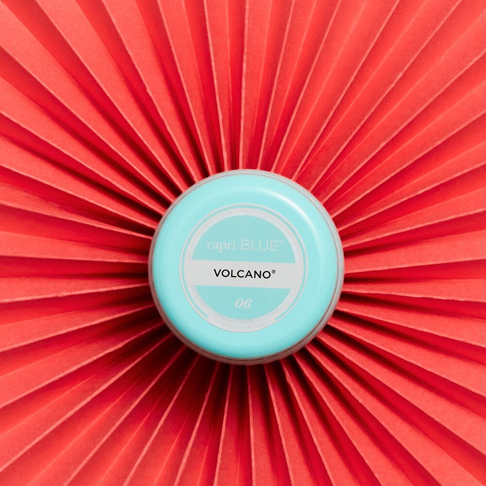 A round, aqua container of Capri BLUE - Volcano 3oz Mini Tin Candle sits at the center of a vibrant red background with symmetrical folds. The Volcano scent, infused with sugared citrus and tropical fruits, enhances the scene with its enticing aroma.