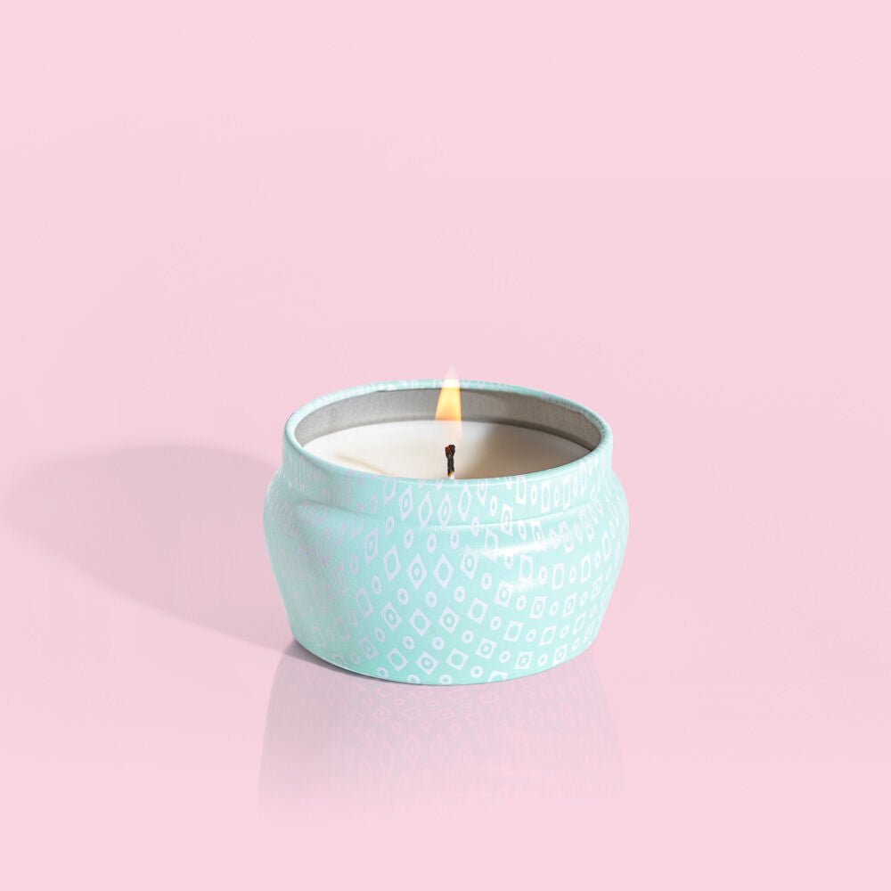 A Capri BLUE - Volcano 3oz Aqua Mini Tin Candle rests in a small, round, light blue tin adorned with a white diamond pattern. Its sugared citrus scent fills the air as the flame glows softly against a pastel pink backdrop, highlighting the candles warmth and elegance.