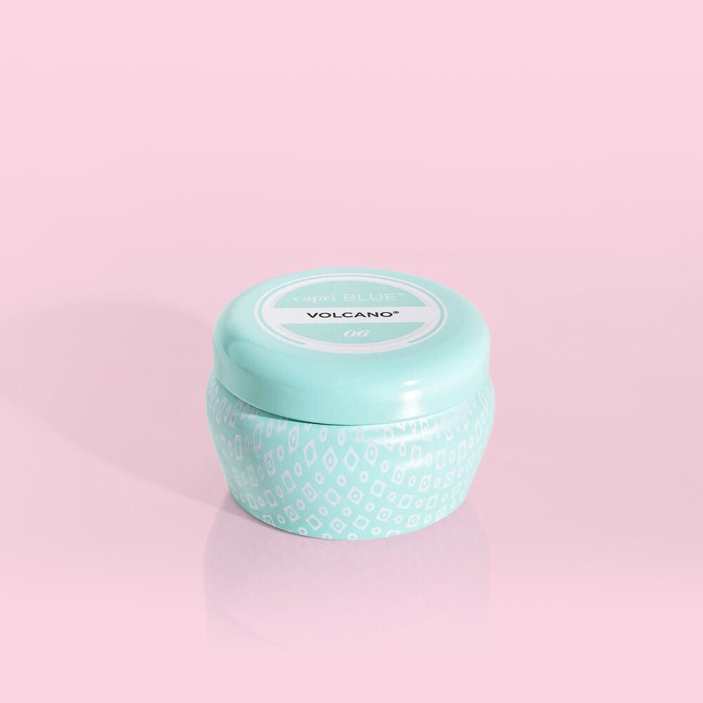 The Capri BLUE - Volcano 3oz Aqua Mini Tin Candle, featuring a decorative teal container and lid, emits the signature Volcano fragrance of sugared citrus. It elegantly sits against a soft pink background.