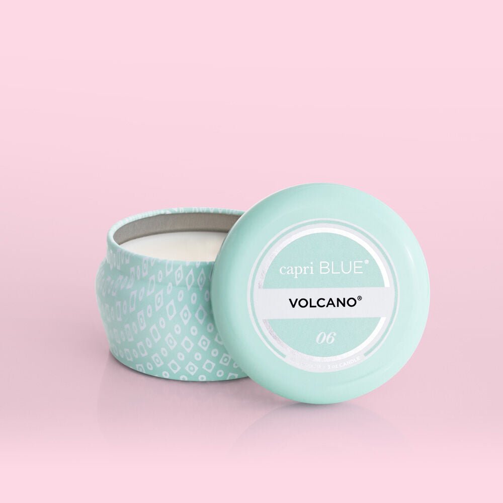 The Capri BLUE - Volcano 3oz Aqua Mini Tin Candle, with its lid off and set on a soft pink background, showcases a white geometric pattern and captures the essence of sugared citrus and tropical fruits in its signature volcano fragrance.